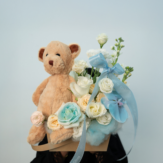 A floral arrangement with a cut phalaenopsis orchid, white roses painted blue, lisianthus, and a teddy bear, all arranged in a rustic kraft box.